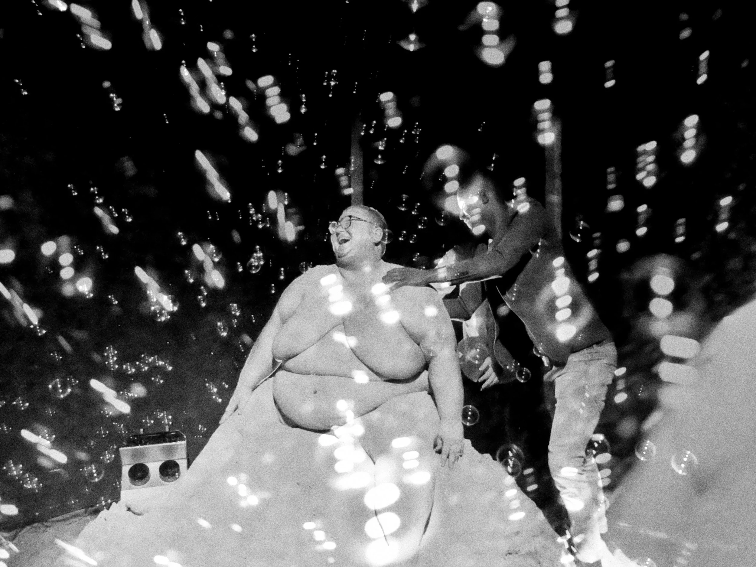  A black and white image of Goldendean. They are naked, and a person is standing beside them touching their shoulder. Goldendean is smiling, and around them the air is filled with bubbles. 