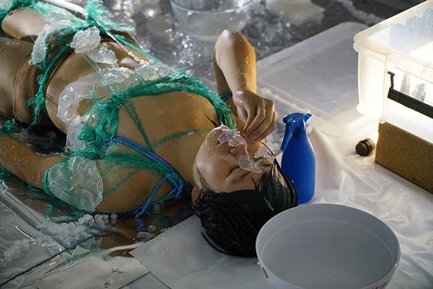  Clarinda is lying on their back. A bowl is by their head and beside them a blue spray bottle. They are wearing an outfit made of green and blue fishing rope and netting. Placed on their body is opaque gels. 