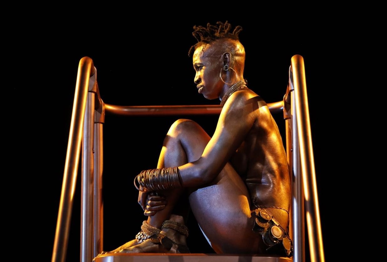  Performer Mamela Nyamza sitting atop a platform, sitting with their knees up held to their body. 