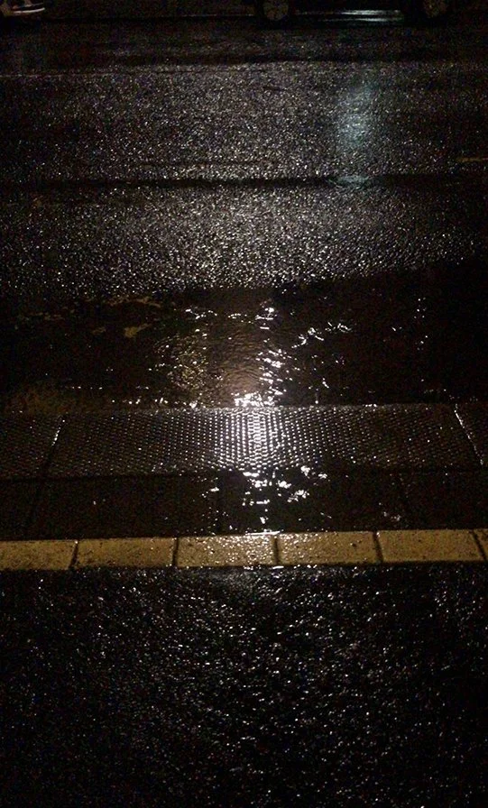  An image of a concrete road that has been rained on, with the reflection of a streetlight in a puddle. Yellow lines cross the road. 