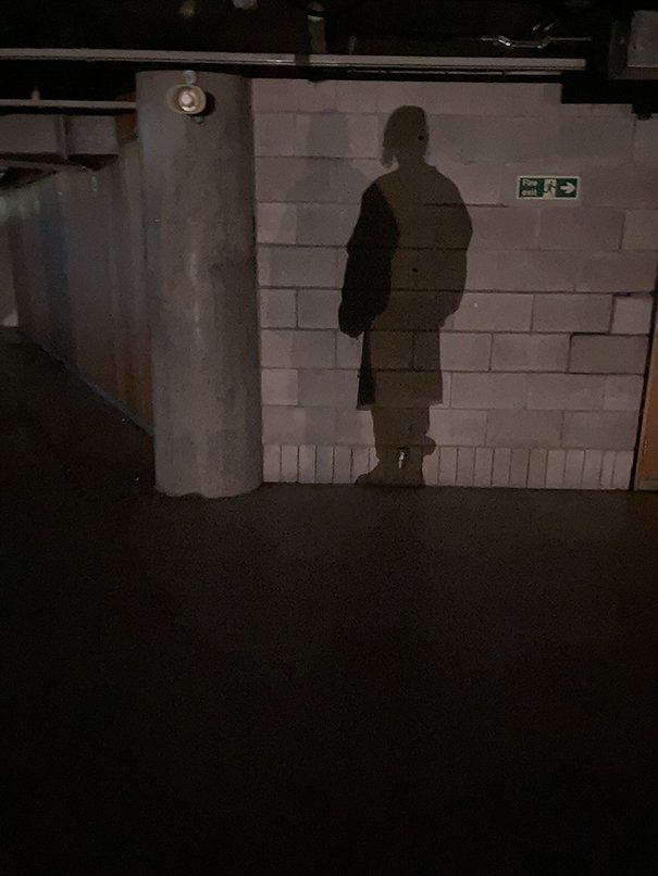  In a dark carpark, NXSA's shadow is in silhouette against a concrete brick wall. There are two overlapping shadows on the wall. In the wall there is also a green fire exit sign. 