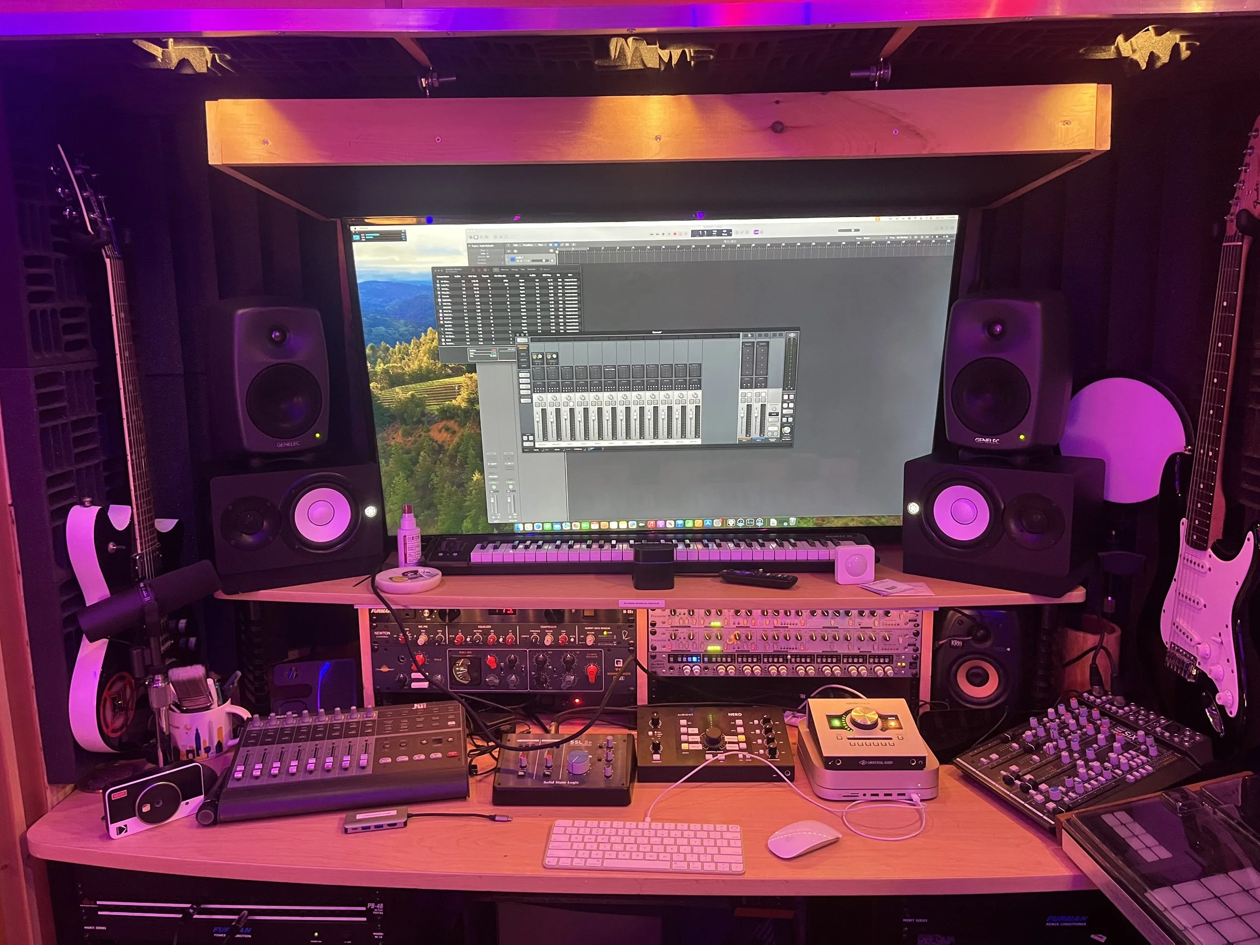 A music production studio setup with a large computer monitor displaying music software, multiple studio speakers, synthesizers, mixers, guitars, and various electronic equipment, all illuminated with purple and pink lighting.
