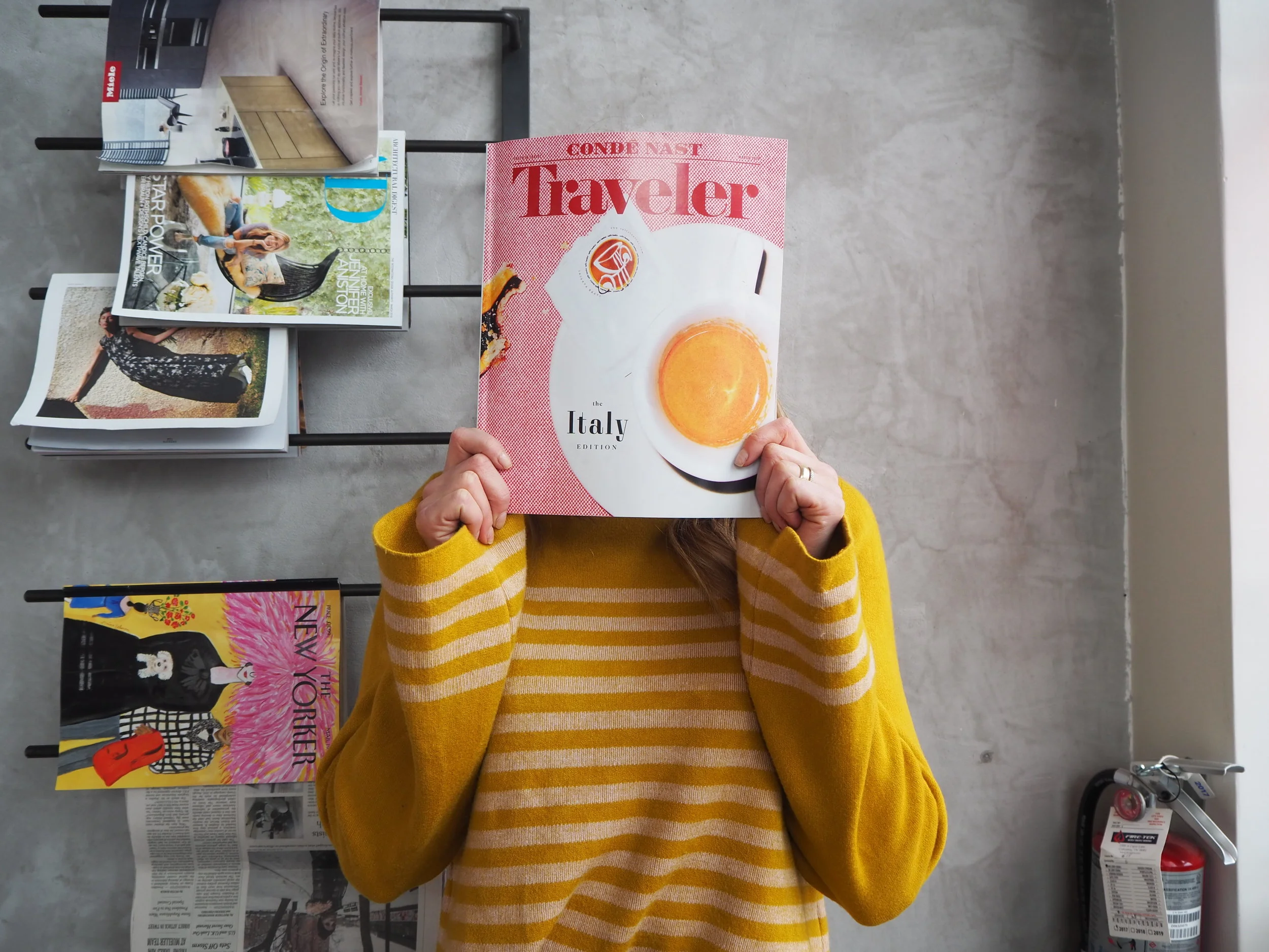 Person in a yellow sweater holding a travel magazine in front of their face, with a background of magazines on a rack.
