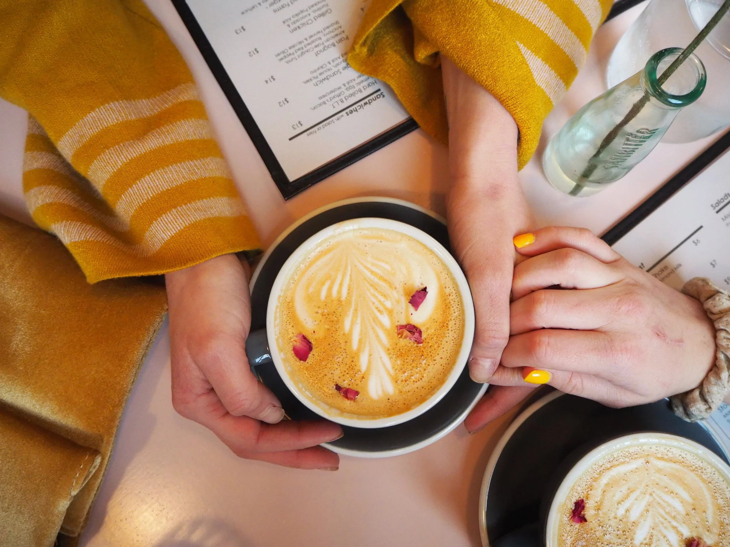 Two people holding hands over a table with latte art coffees, wearing yellow-striped sweaters and a yellow painted nail.