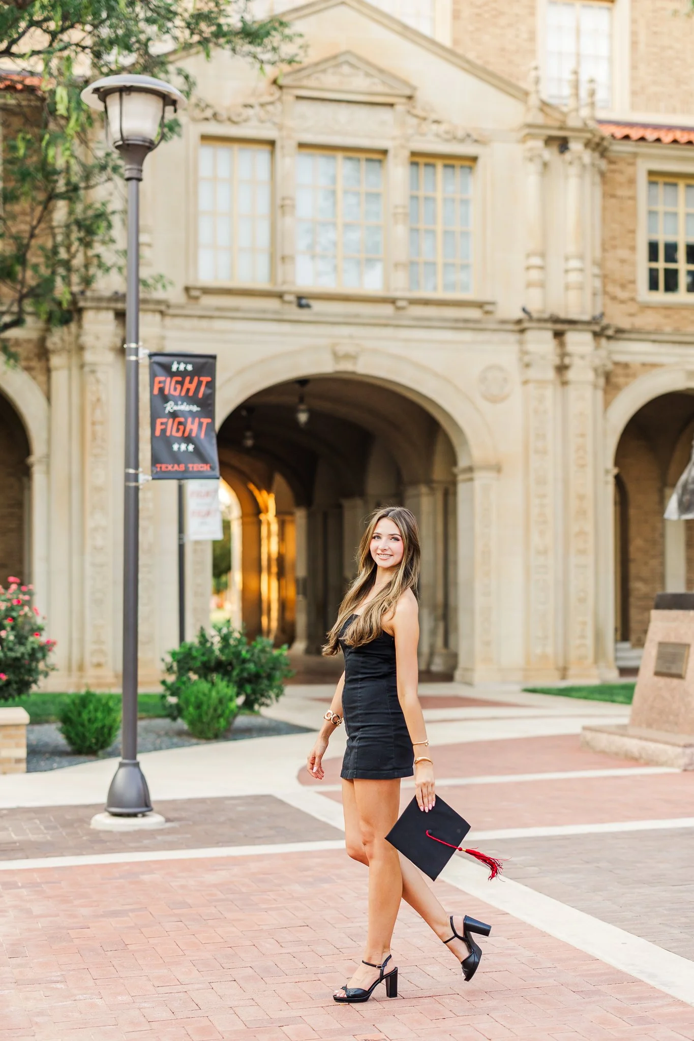 college girl from texas tech is walking with cap on her hand while wearing a black dress. she is posing and standing in the admin building of texas tech campus
