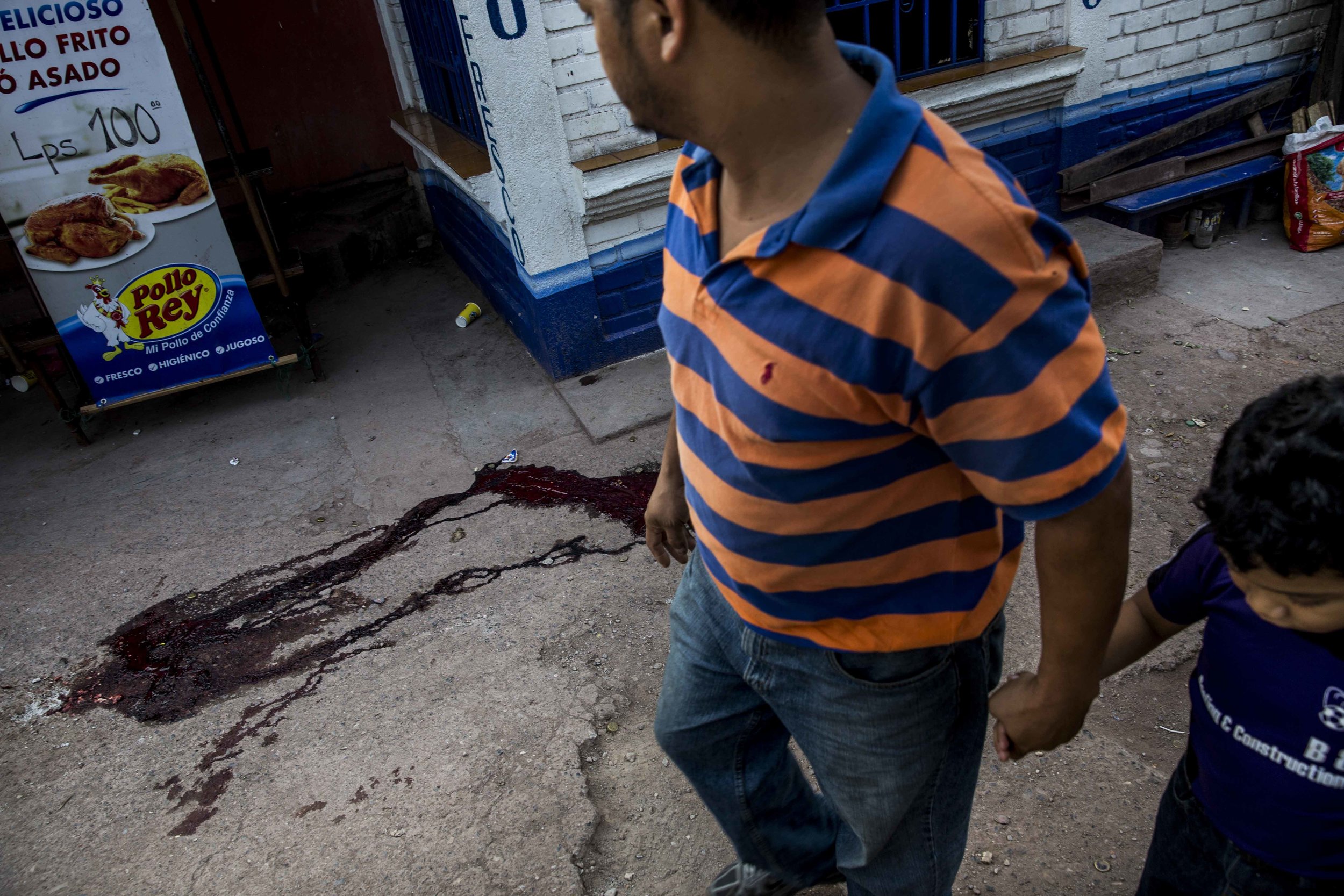 Father and son walk by next to the pool of blood left by the dead body of Edwin Amador, 20, murdered in cold blood in the streets of the Suyapa district, Teguciaglpa, capital of Honduras.