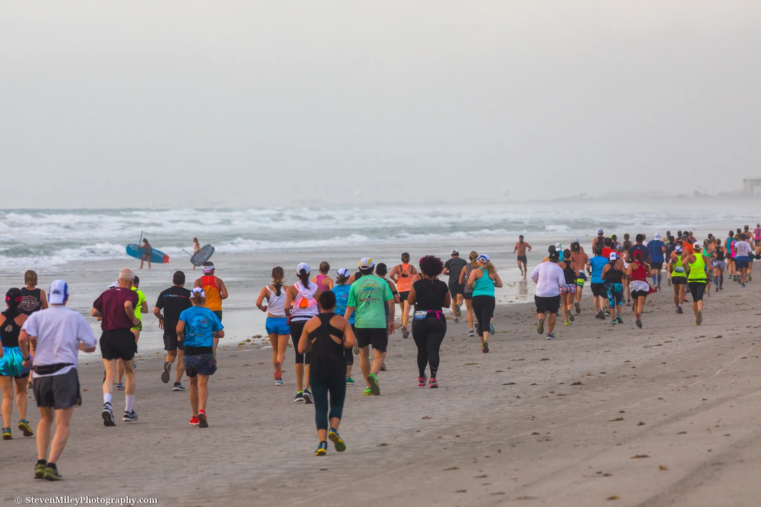 A pair of paddleboarders sneak through a long line of runners participating in the U.S.A. Beach Running Championships. Cocoa Beach, FL.