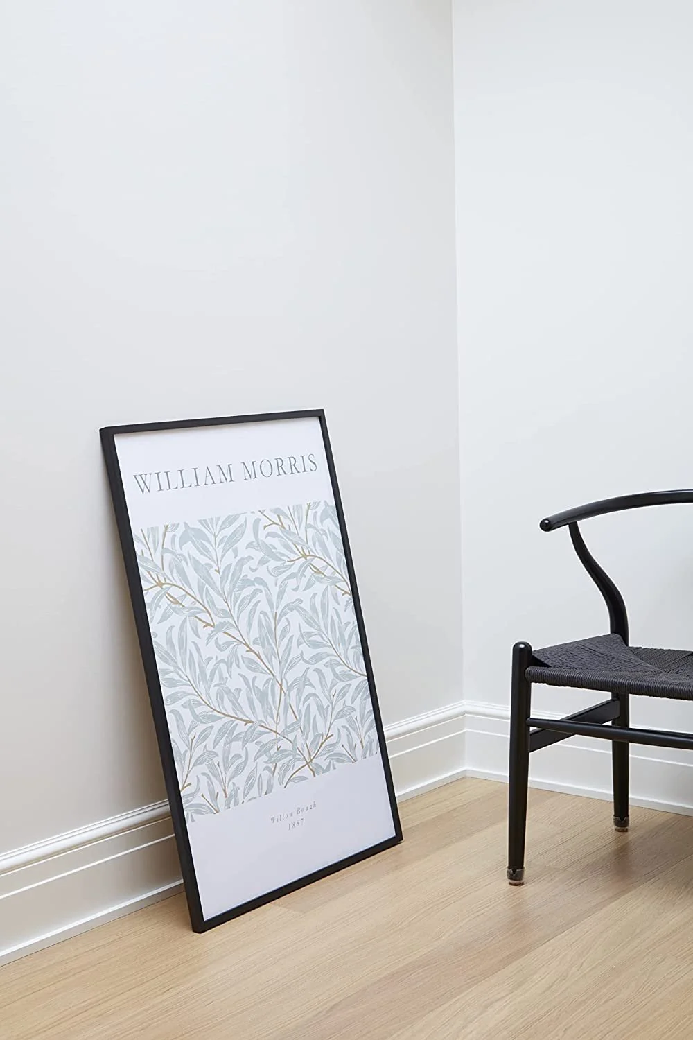 Framed William Morris art print leaning against a white wall next to a black wooden chair with woven seat on a light wood floor.