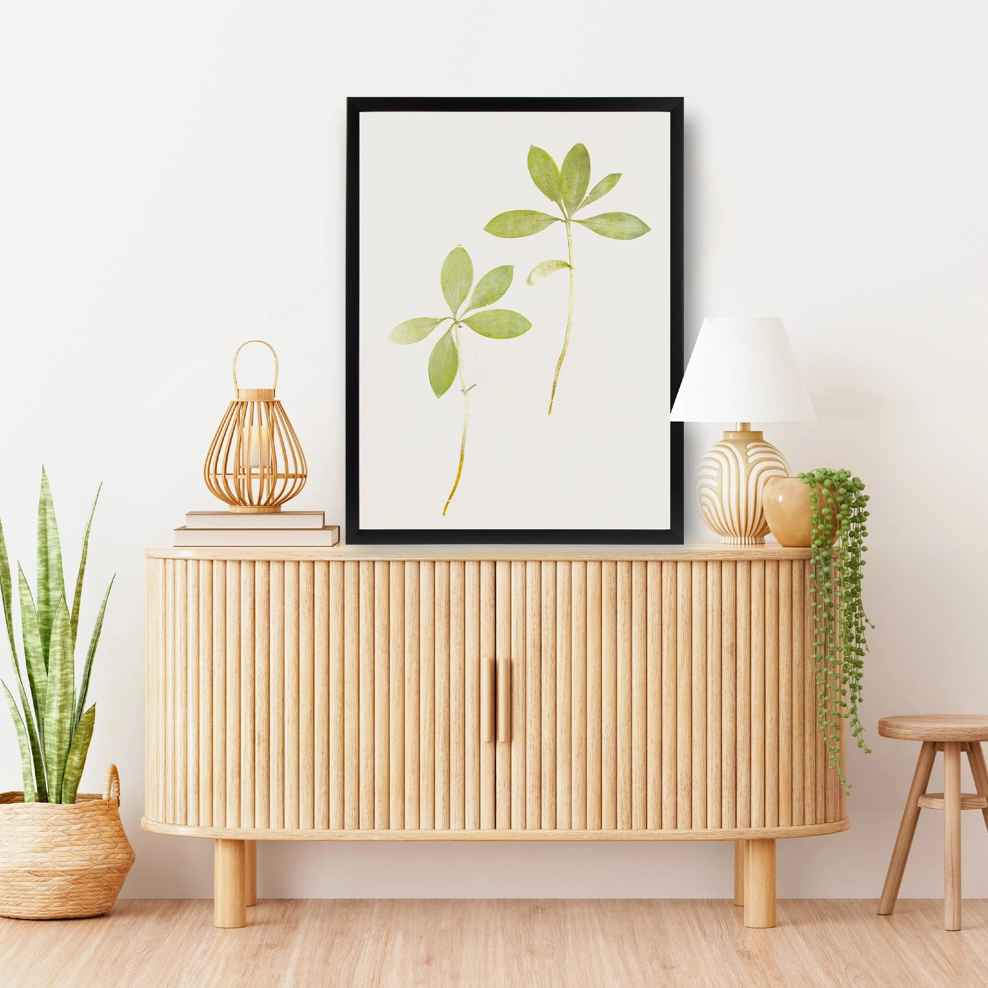 Decorative sideboard with botanical artwork, a table lamp, a wicker lantern, and potted plants, set against a white wall.