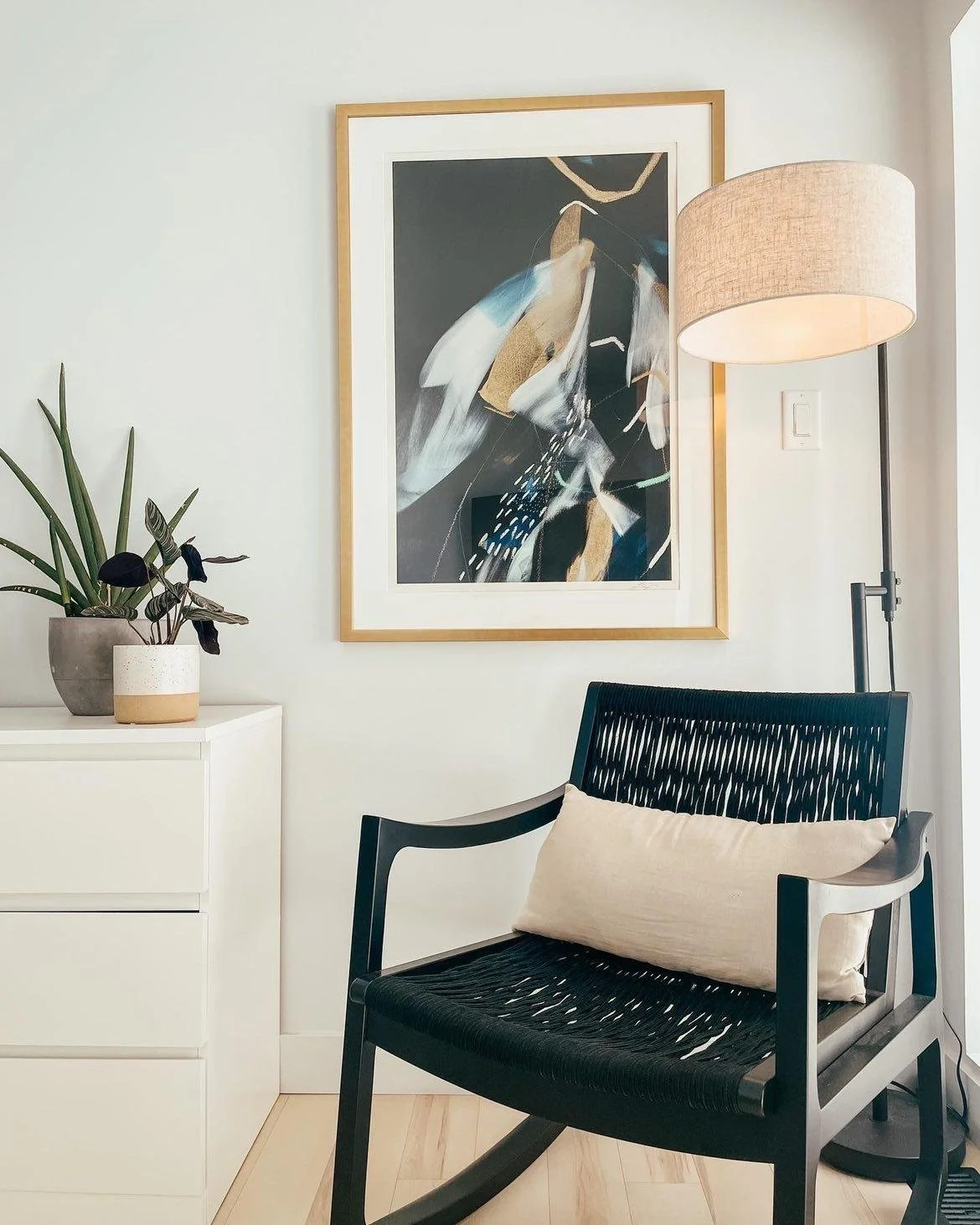 A modern interior corner with a black woven chair featuring a beige pillow, a white dresser with two potted plants, a large abstract artwork on the wall, and a tall floor lamp with a beige shade.