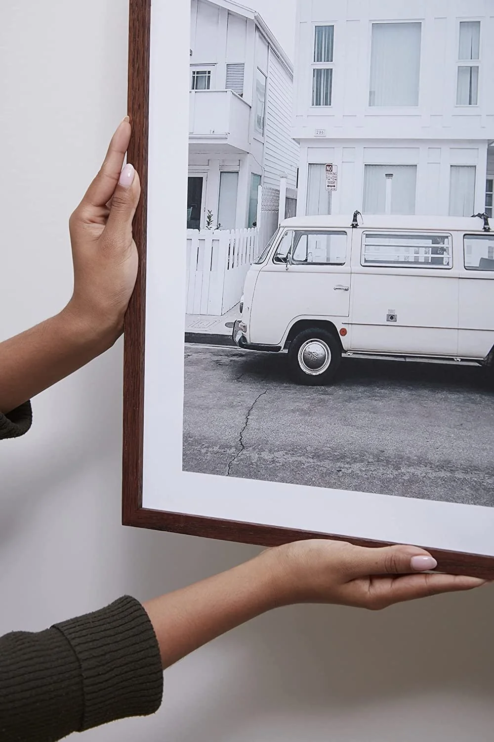 Person holding a framed black-and-white photo of a white vintage van parked on a street in front of a white building and fence.