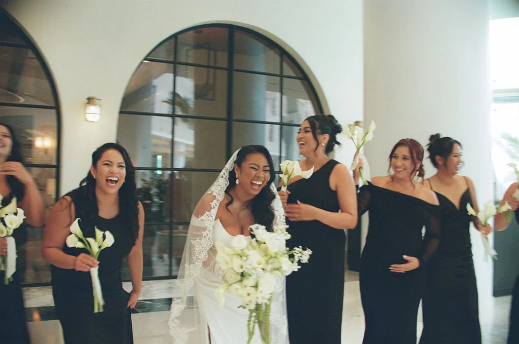 A bride in a white wedding dress and veil, holding a bouquet of white flowers, is surrounded by bridesmaids in black dresses, all smiling and holding white flowers, in a bright, modern venue with arched windows.