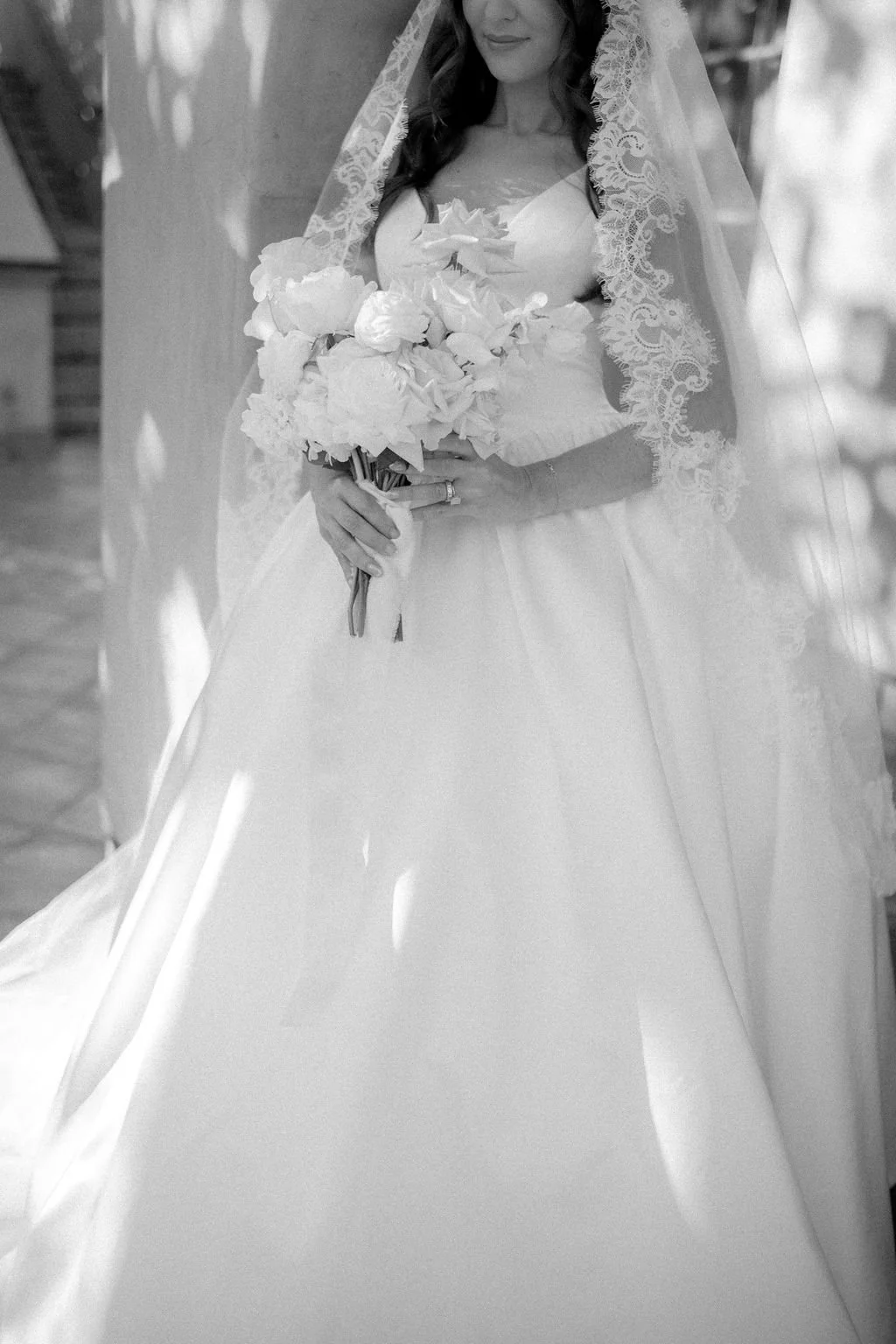 A bride in a wedding gown holding a bouquet of flowers, with a lace veil over her head, standing indoors.