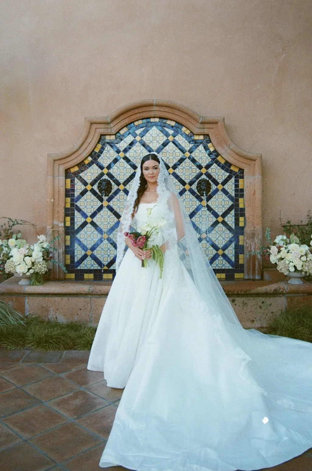 A bride in a white wedding gown holding a bouquet of flowers, standing in front of a decorative tiled fountain with floral arrangements on either side.