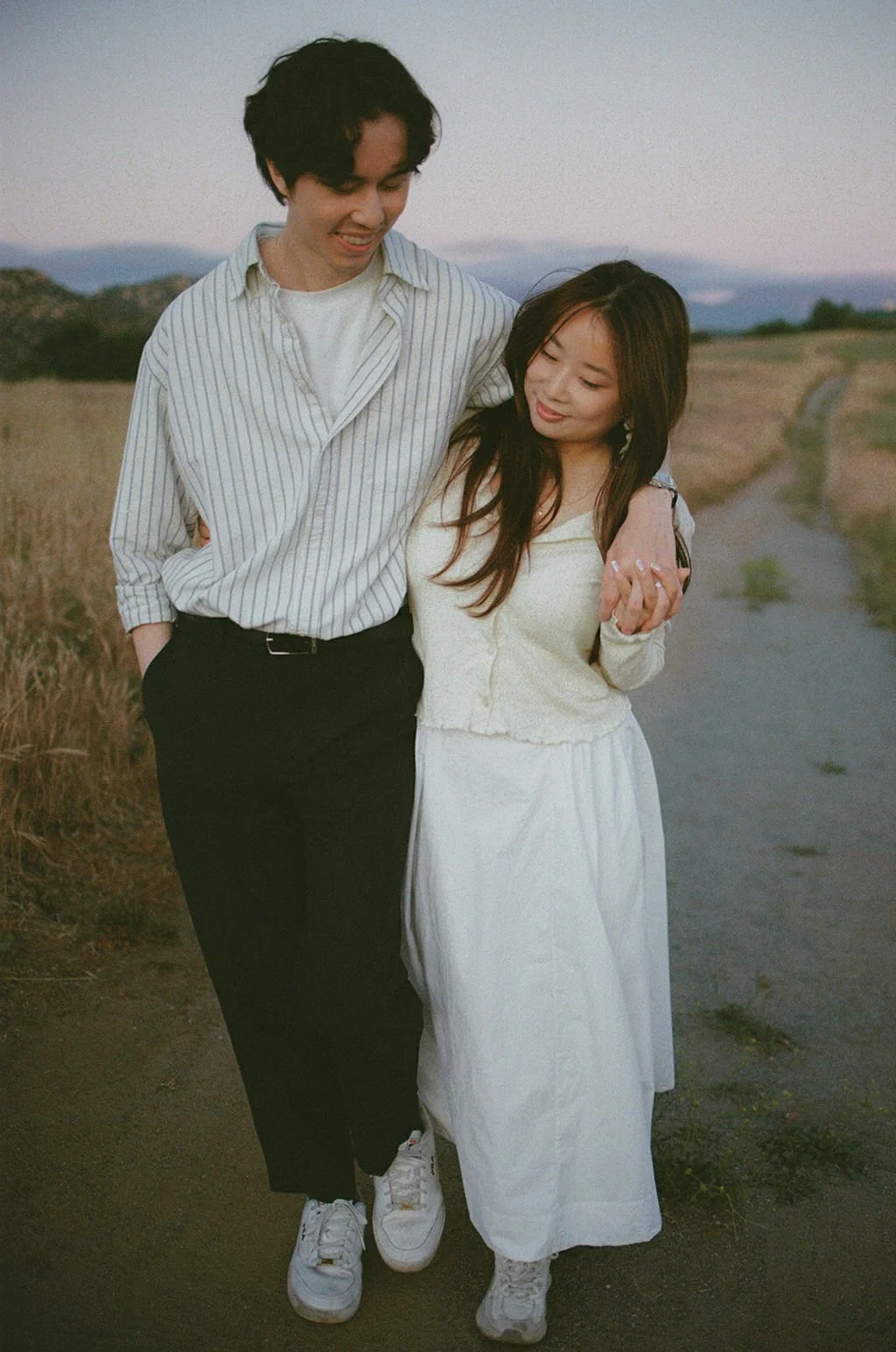 A young couple walking together on a rural path during sunset, smiling and holding hands while looking at each other.