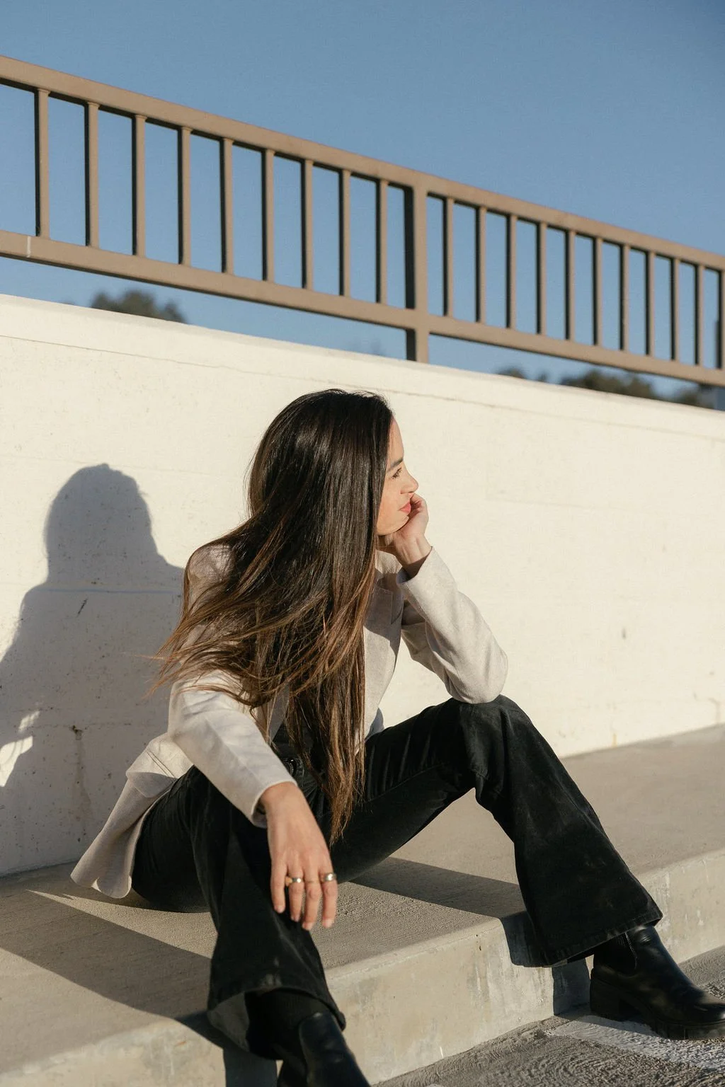 A woman sitting on a concrete bench, looking to her right with her hand supporting her head. She has long brown hair, wears a light-colored blazer, black pants, and black boots. The sun casts a shadow of her profile on the white wall behind her, with a clear blue sky and a beige railing above.