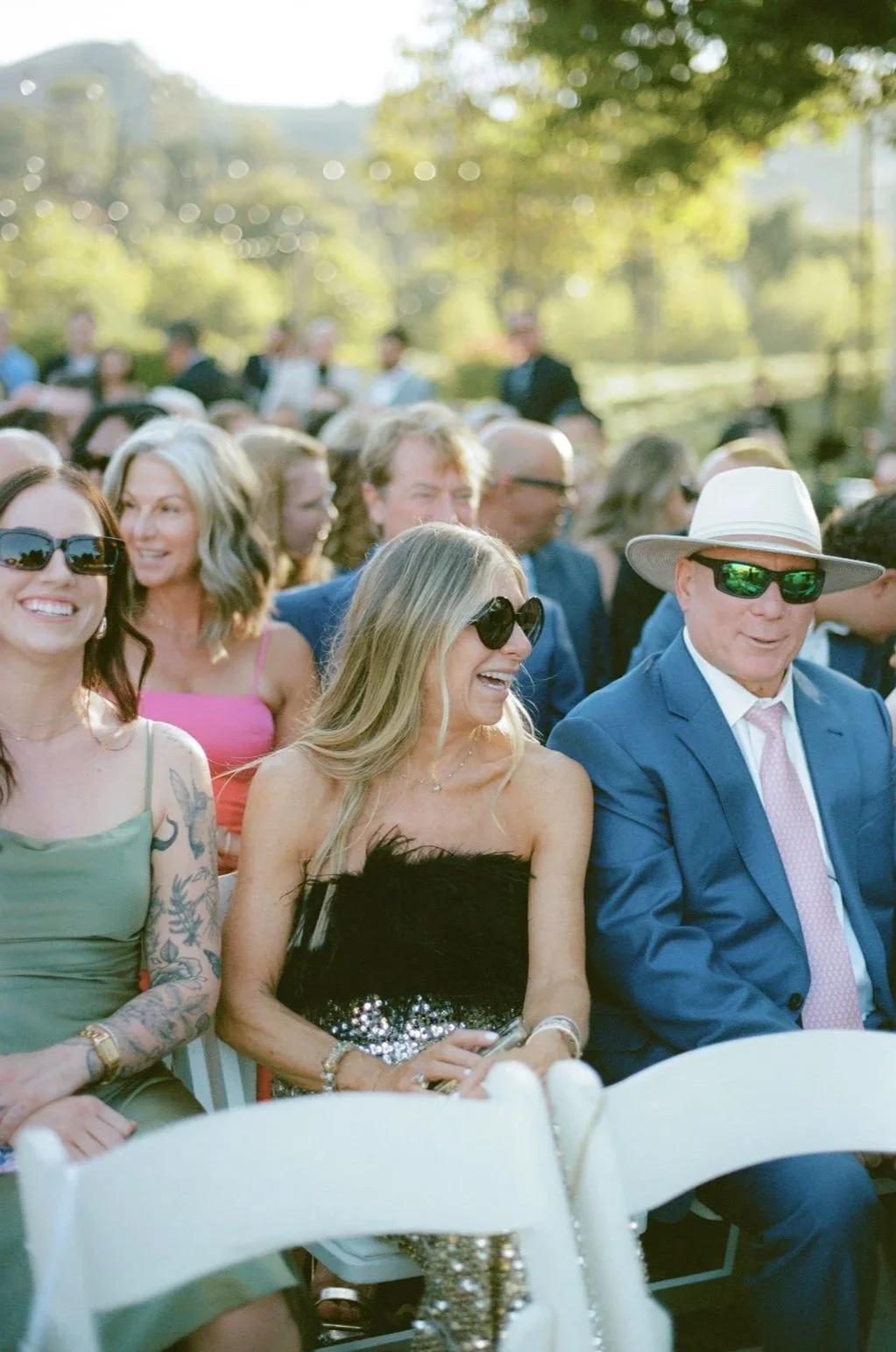 People sitting and smiling at an outdoor event, with a woman in a black strapless dress and sunglasses, a man in a blue suit with a pink tie and white hat, and others dressed formally, during daytime.