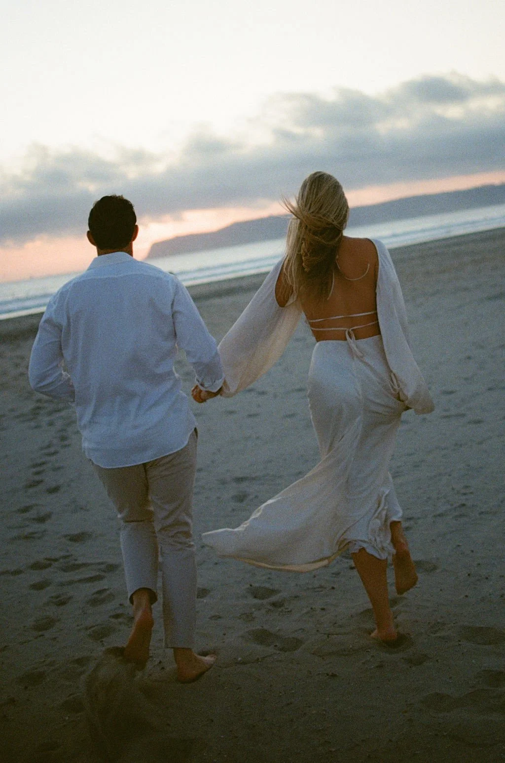 A couple walking hand in hand on a beach at sunset, wearing white clothing.