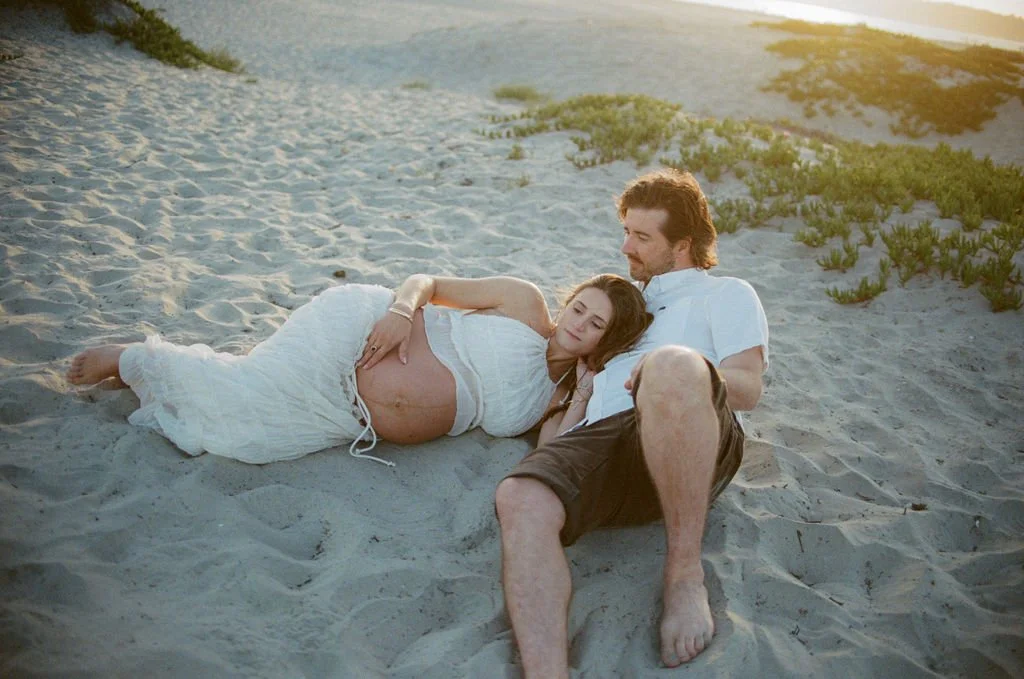 A couple sitting on a sandy beach at sunset, the woman is pregnant and resting her head on the man's shoulder, both are casually dressed and looking peacefully into the distance.