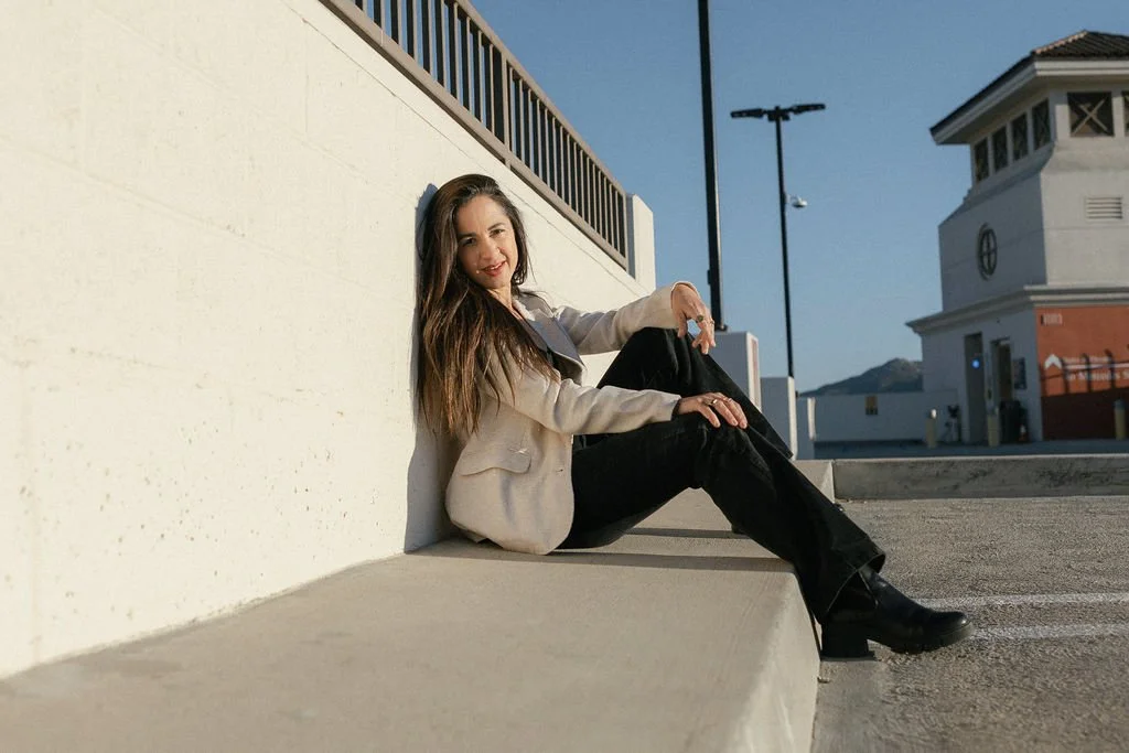 A woman sitting on the ground leaning against a white wall outdoors on a sunny day.