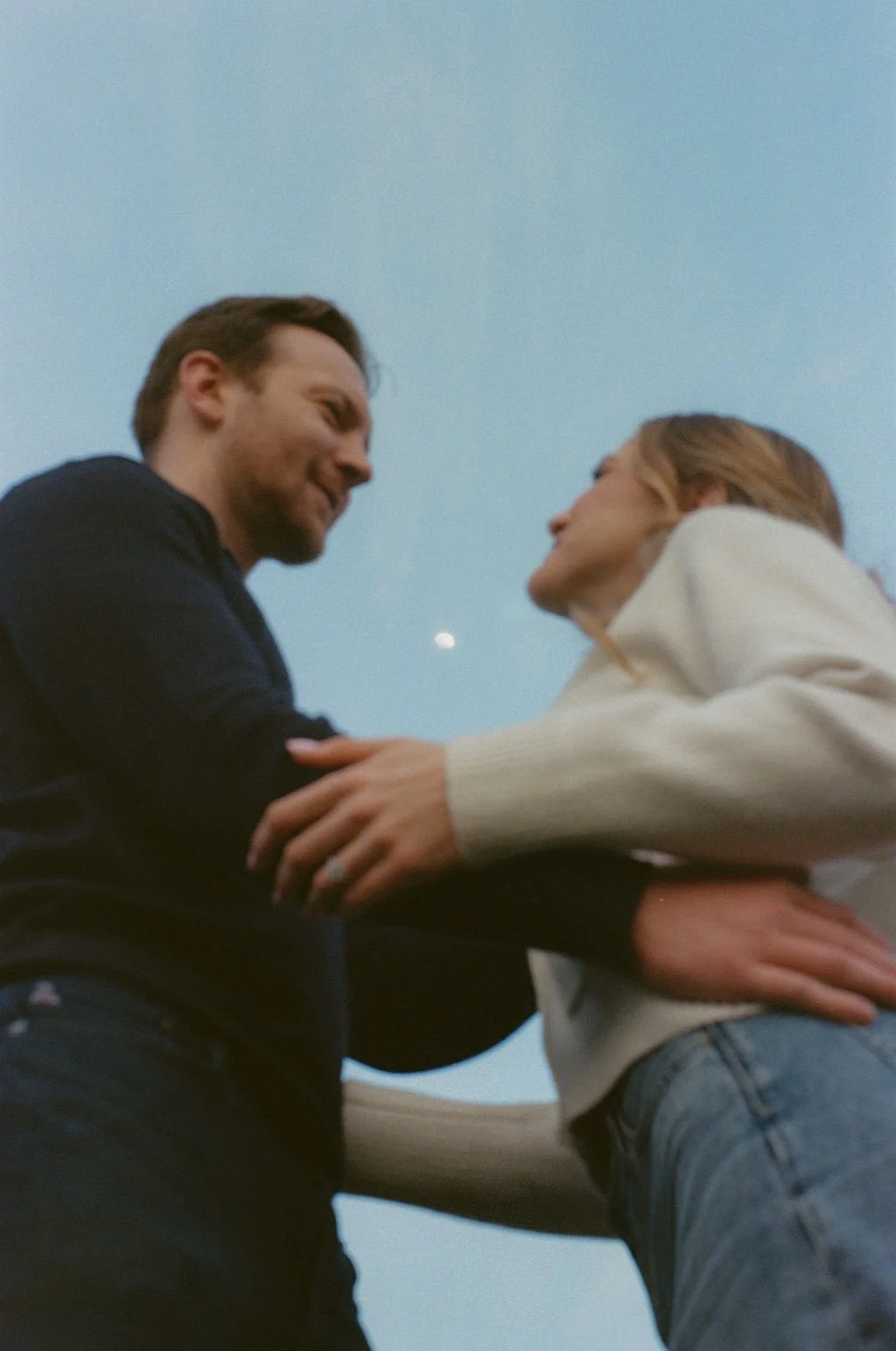 A couple looks into each other's eyes while holding hands and leaning on a piece of furniture outdoors during dusk.
