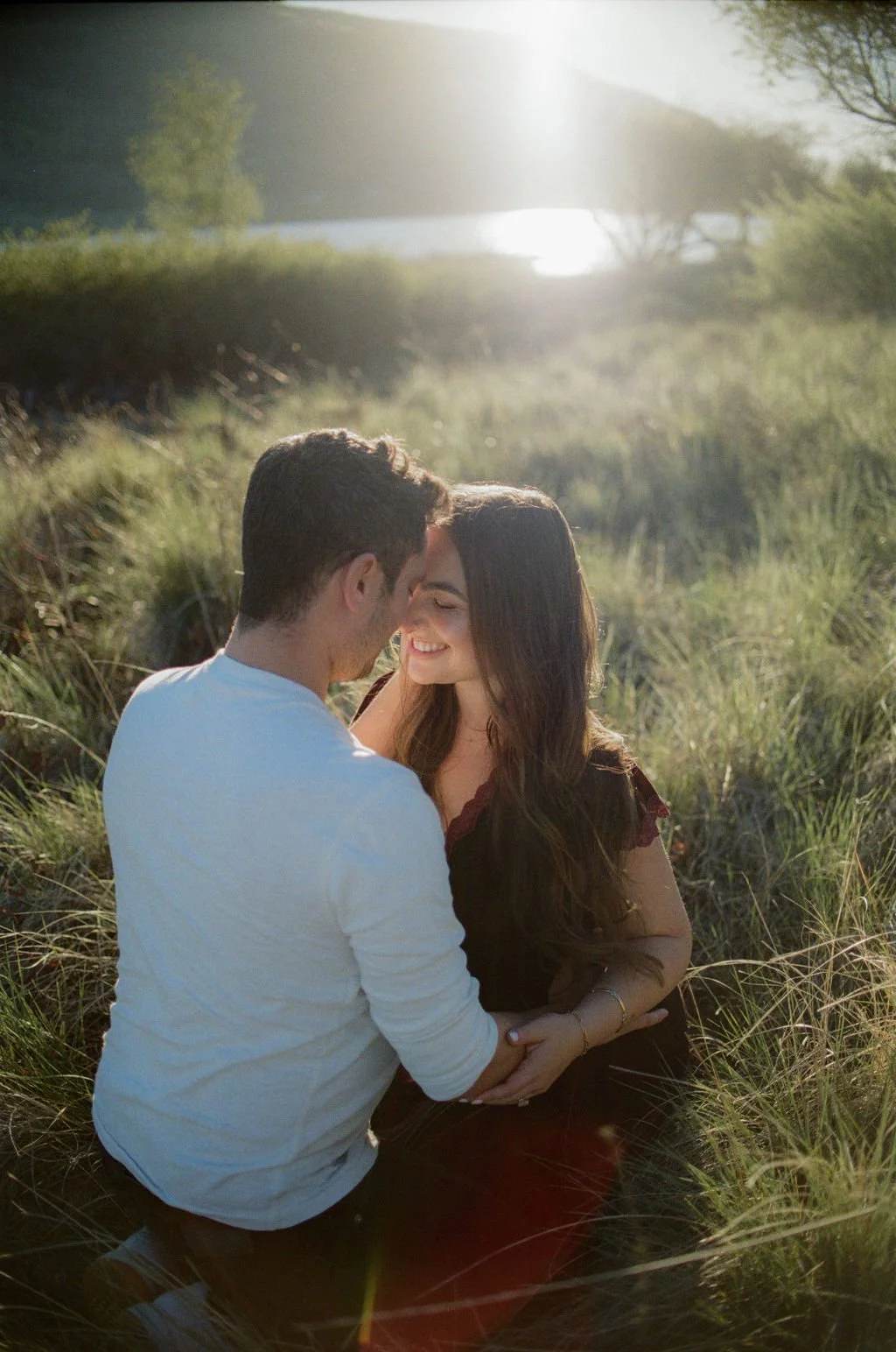 A couple sitting closely together in a grassy field during sunset, smiling and sharing an intimate moment.