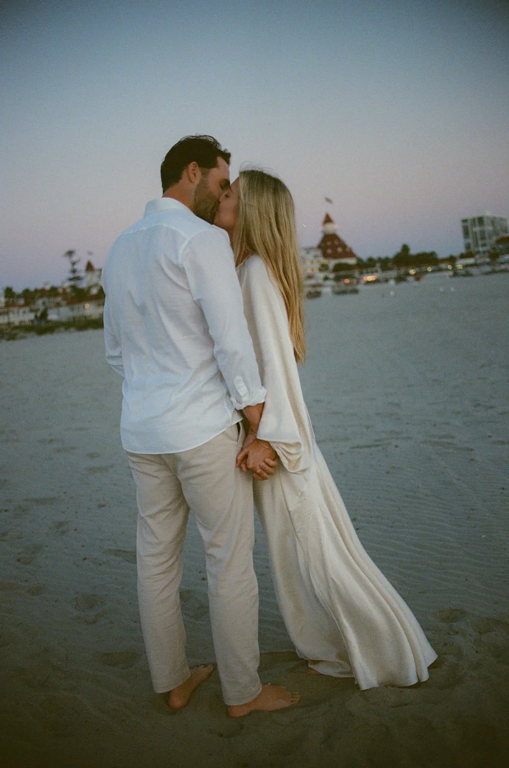 A couple sharing a kiss on a beach at sunset, holding hands, with buildings and boats in the background.