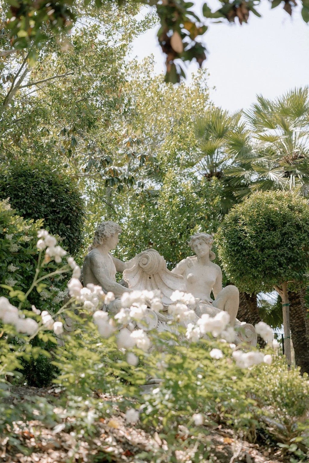 A classical marble sculpture of a man and woman surrounded by lush greenery including trees and flowering bushes.