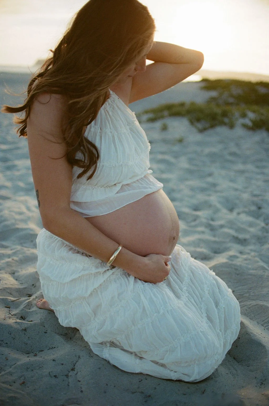 Pregnant woman sitting on the beach during sunset, wearing a white dress and holding her belly.