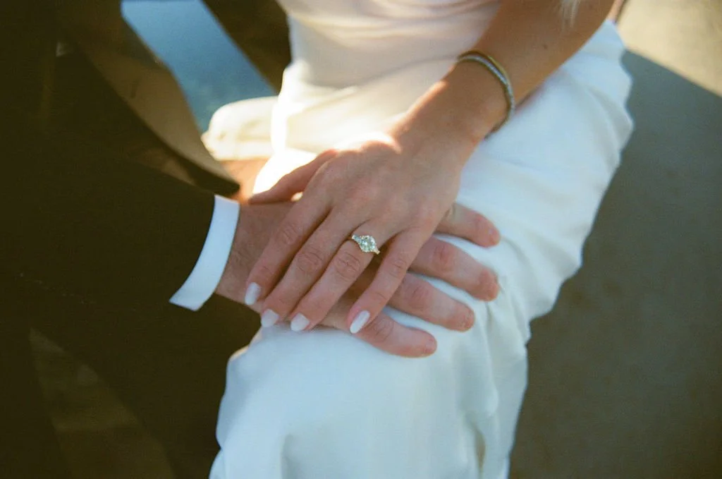 Close-up of a couple's hands, showing a wedding ring on the woman's finger, with the bride wearing a white outfit and the groom in a dark suit.