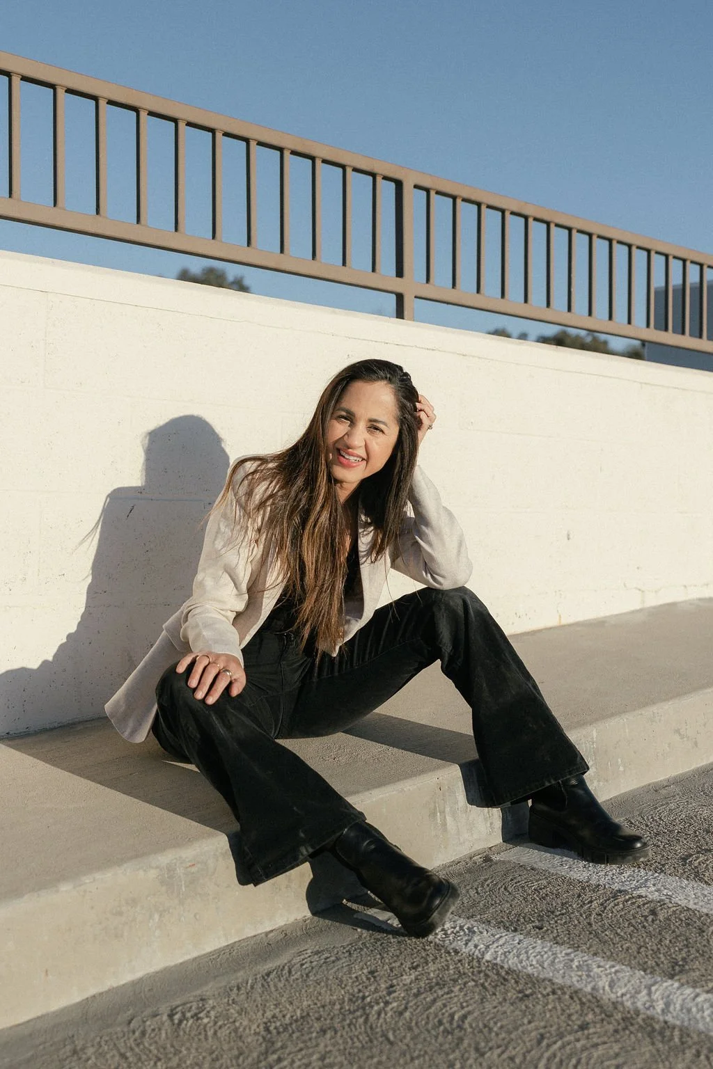 A woman with long brown hair sitting on a curb, smiling, dressed in a beige blazer, black pants, and black boots under a blue sky.