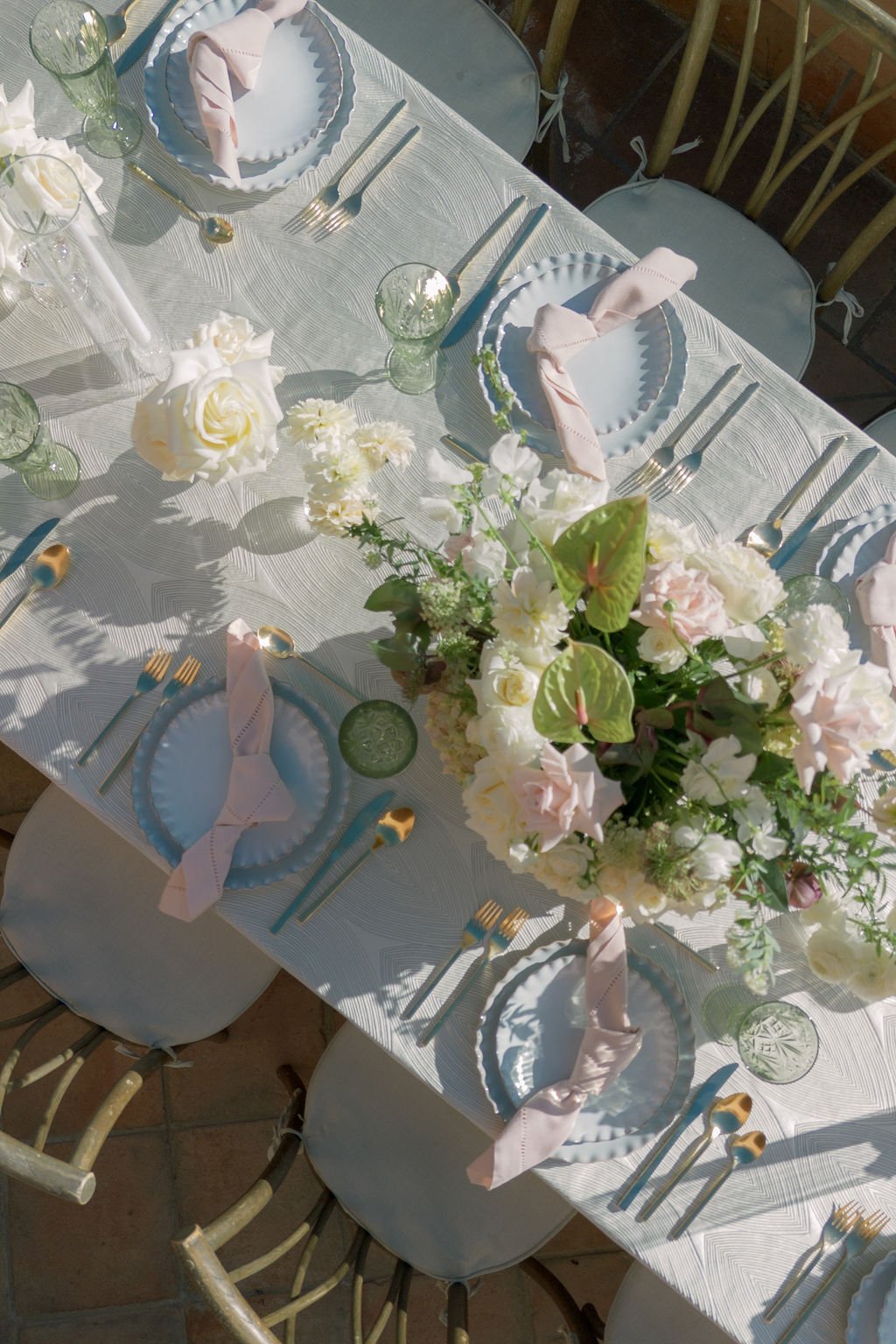 A dining table set for a formal event with white plates, pink napkins, gold utensils, clear drinking glasses, and a floral centerpiece with white and pink flowers and green leaves.