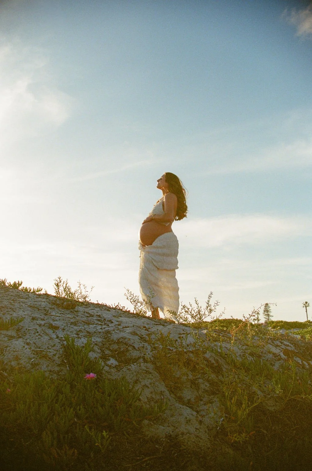 A pregnant woman standing outdoors on a low hill during sunset, wearing a white dress, with her hands resting on her belly and looking up, with a few plants and a tree in the background.