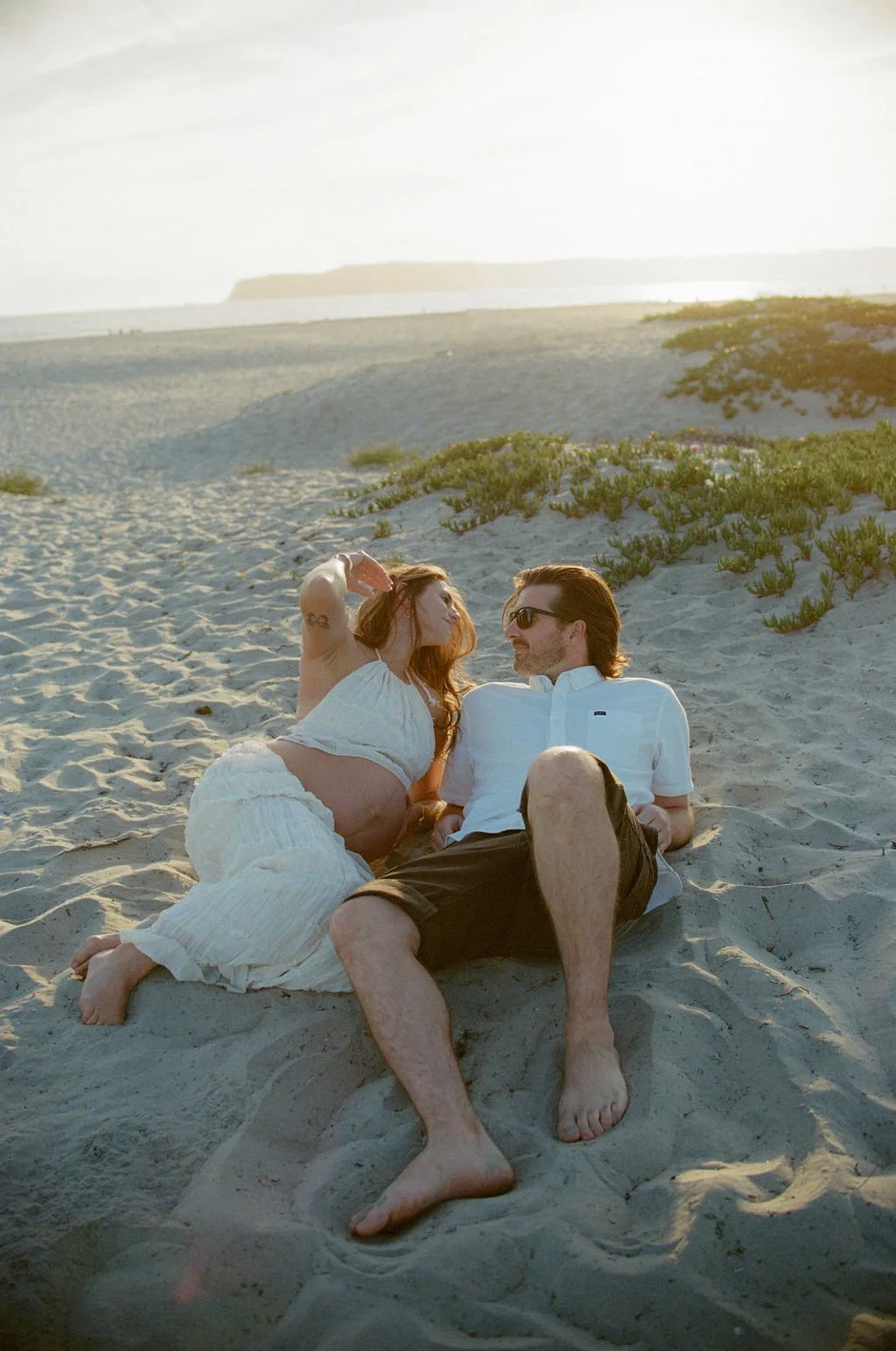 A couple lying on the sand at the beach, gazing at each other, with one woman touching her hair and the man wearing sunglasses. The sun is setting in the background, creating a warm glow.