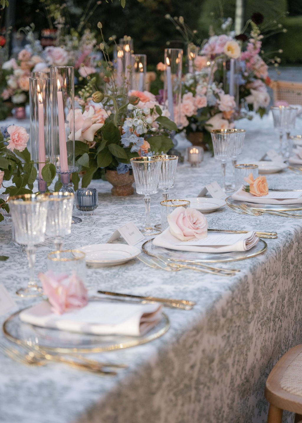 Elegant outdoor dining table decorated with pink and white roses, greenery, tall candles, and fine glassware. The table has white plates with gold accents, napkins with pink roses, and a textured tablecloth.