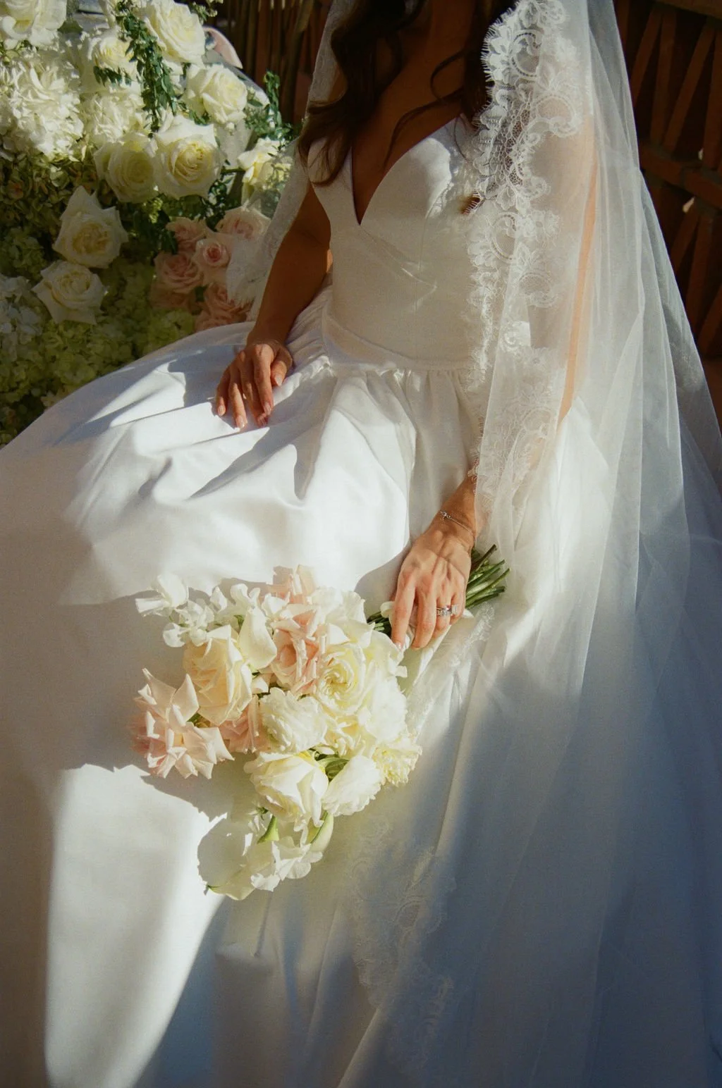 A bride in a white wedding gown with lace details, holding a bouquet of white and pale pink flowers, sitting near a large floral arrangement of white roses and greenery.