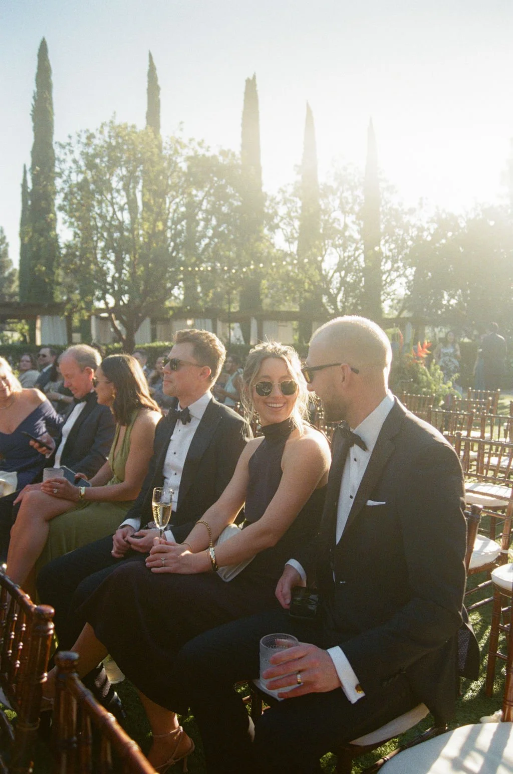 People dressed in formal attire attending an outdoor event during sunset, sitting on chairs, smiling, and holding drinks, with trees and cypress plants in the background.