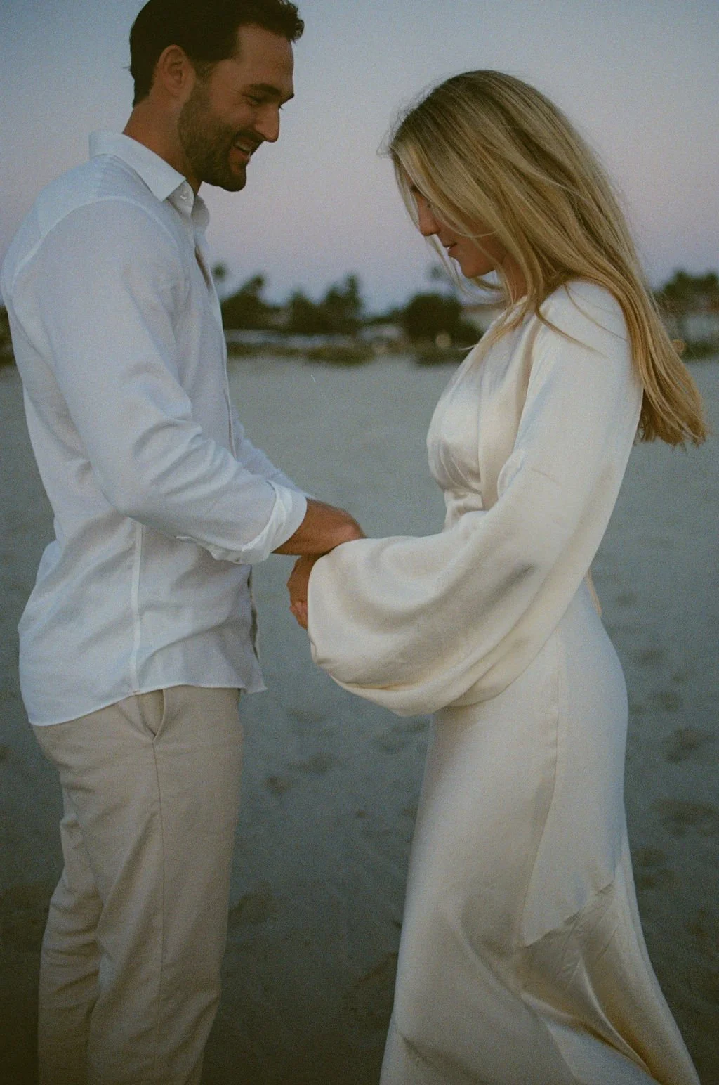 A man and woman holding hands and smiling at each other on a beach during sunset.