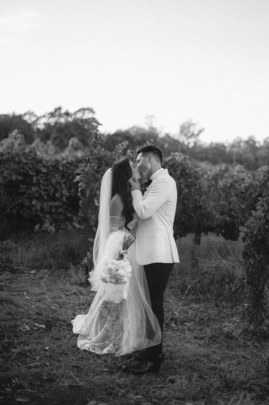A black-and-white photo of a bride and groom in a vineyard, embracing and about to kiss. The bride holds a bouquet and wears a wedding dress with lace details, while the groom is dressed in a white tuxedo jacket and dark pants.
