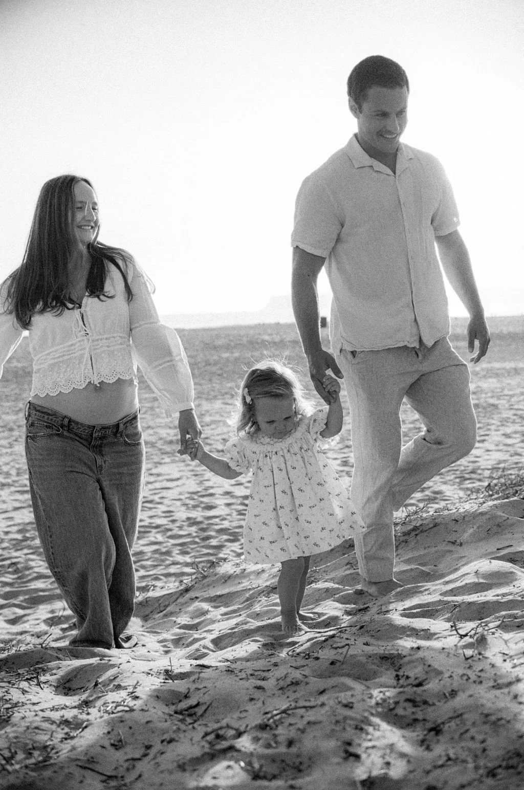 A family of three, including a young girl, walking on a sandy beach holding hands, with the parents smiling as they walk in the late afternoon or evening.