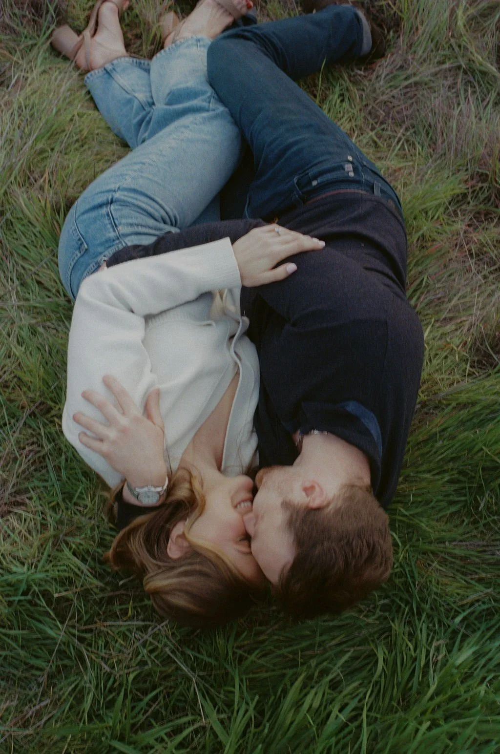 A couple lying on the grass, sharing a kiss and holding each other in a romantic outdoor setting.