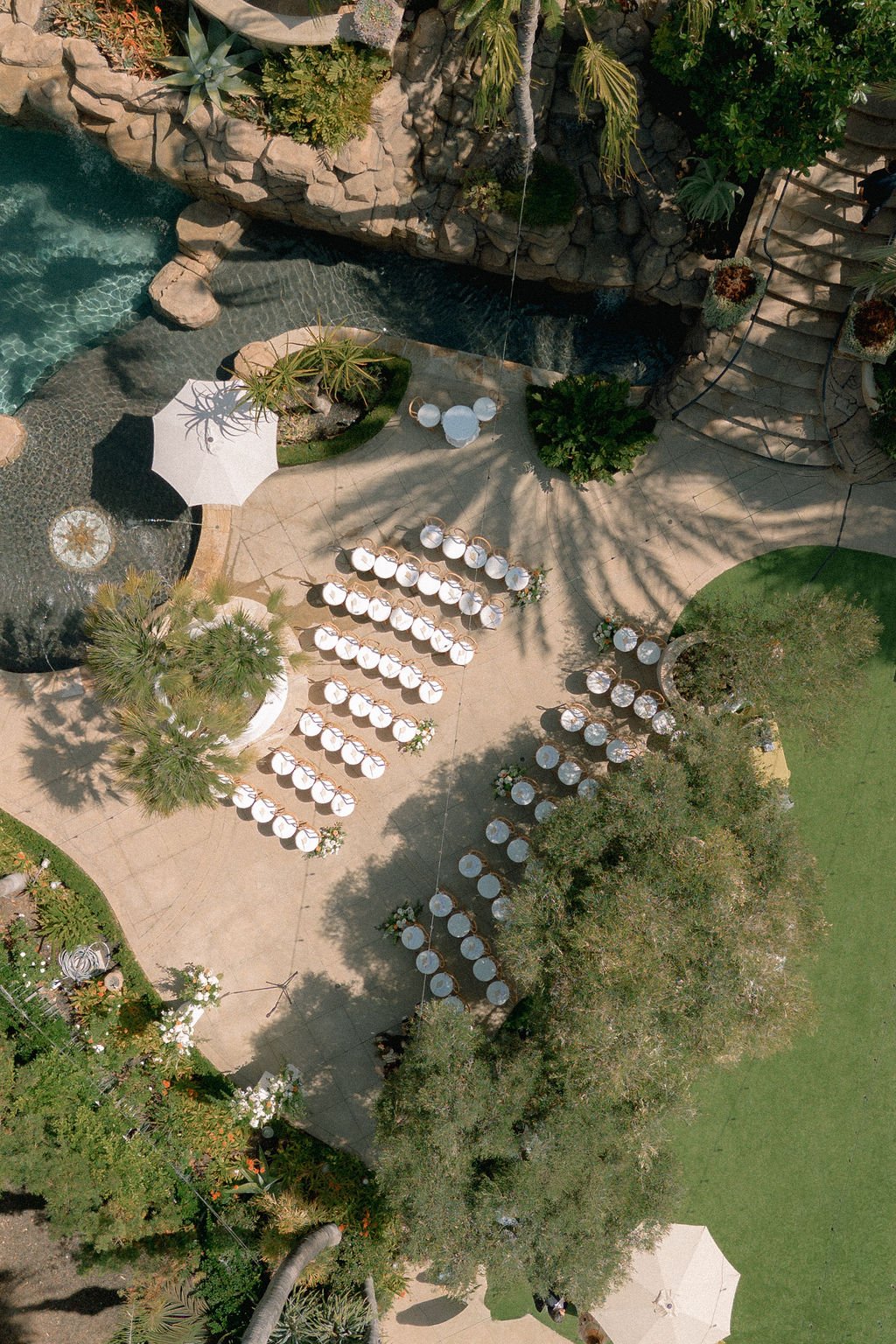 An aerial view of an outdoor area with a swimming pool, a waterfall feature, a shaded dining area with white tables and chairs, and landscaped greenery and trees.