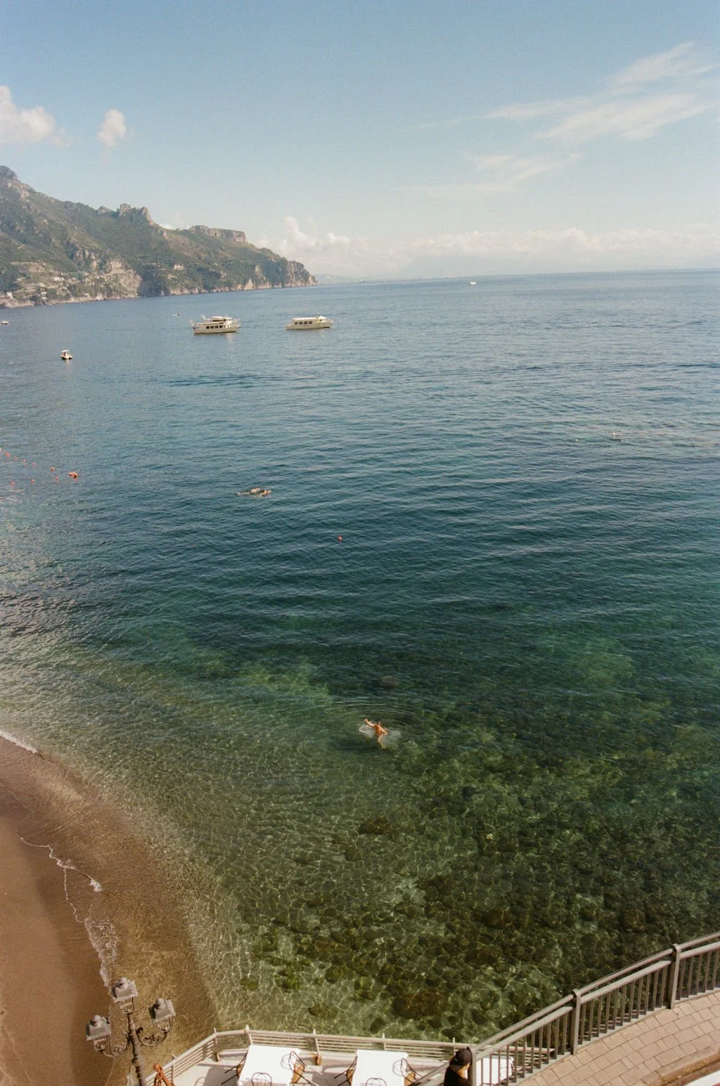 The image shows a scenic view of a calm ocean with two boats floating on the water. A person is swimming near the shore, which has a sandy beach and rocks visible through the clear water. In the background, there are green mountains and a partly cloudy sky.