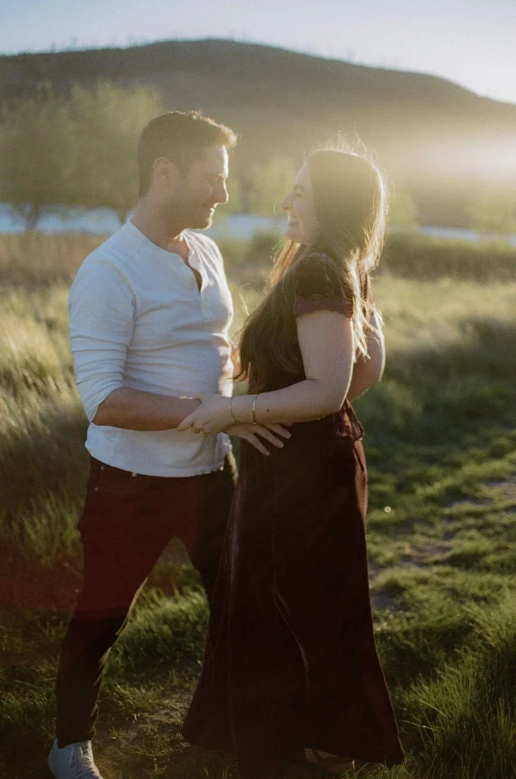 A young couple standing close in a grassy outdoor field during sunset, looking at each other and smiling.