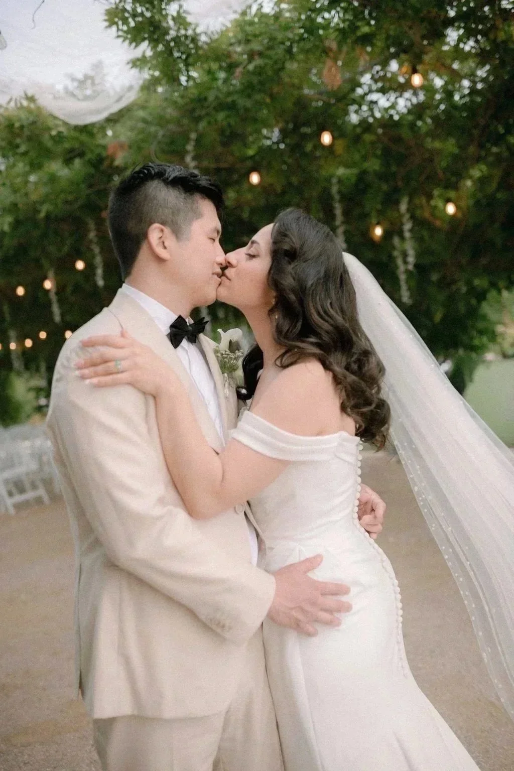 A newlywed couple sharing a kiss outdoors, surrounded by string lights and trees, with the bride wearing a white off-shoulder wedding gown and veil, and the groom in a beige suit with a black bow tie.