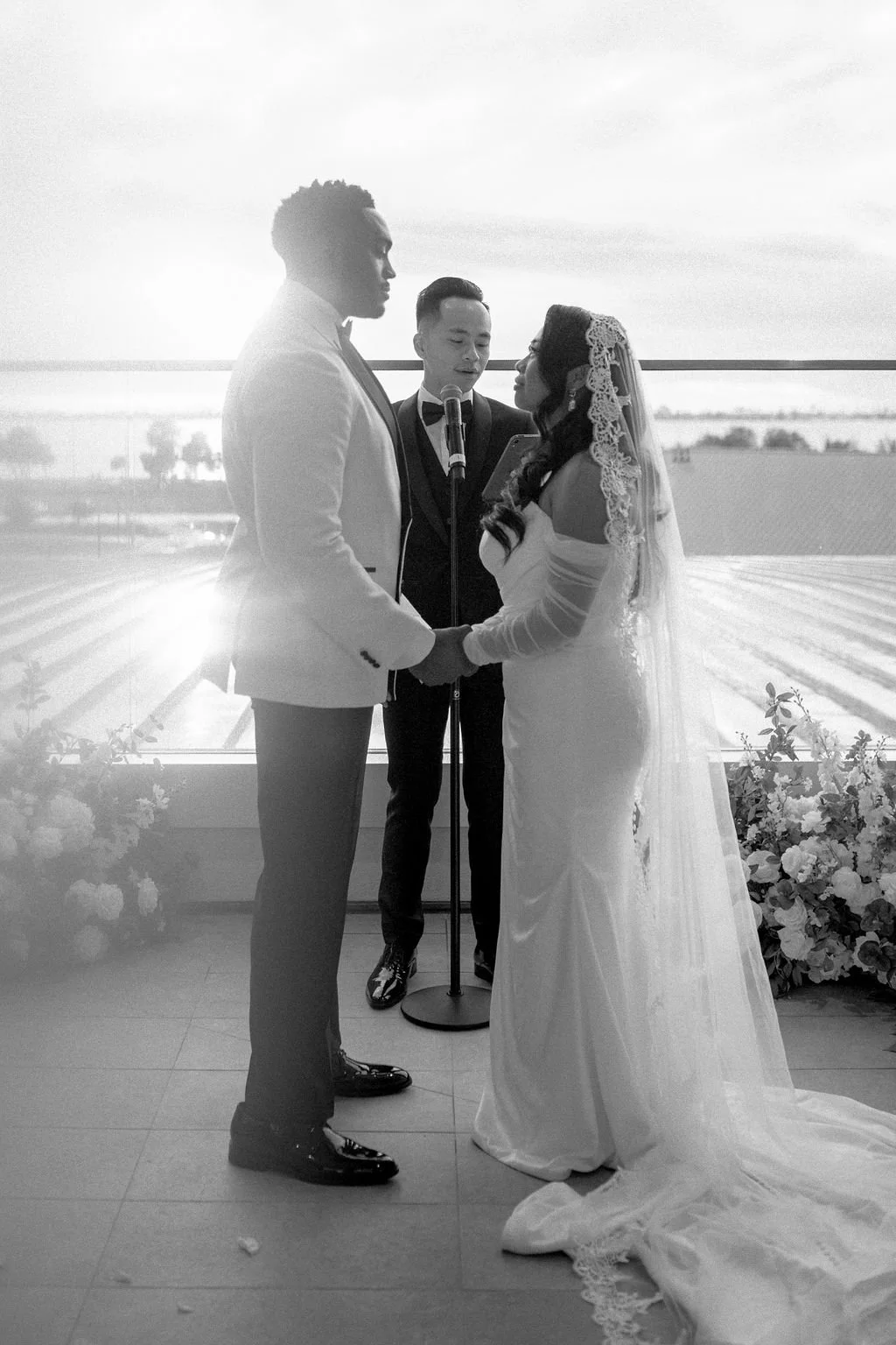 A black and white photo of a couple getting married, holding hands and facing each other, with an officiant standing behind them. The groom is wearing a white tuxedo jacket with black pants, and the bride is in a wedding gown with a veil. They are exchanging vows or rings, with a scenic outdoor view in the background and flowers at their feet.