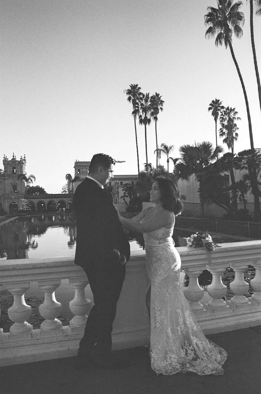 A black and white photo of a bride and groom dancing outdoors on a balcony with a balustrade, overlooking a water feature and palm trees, during sunset or sunrise.