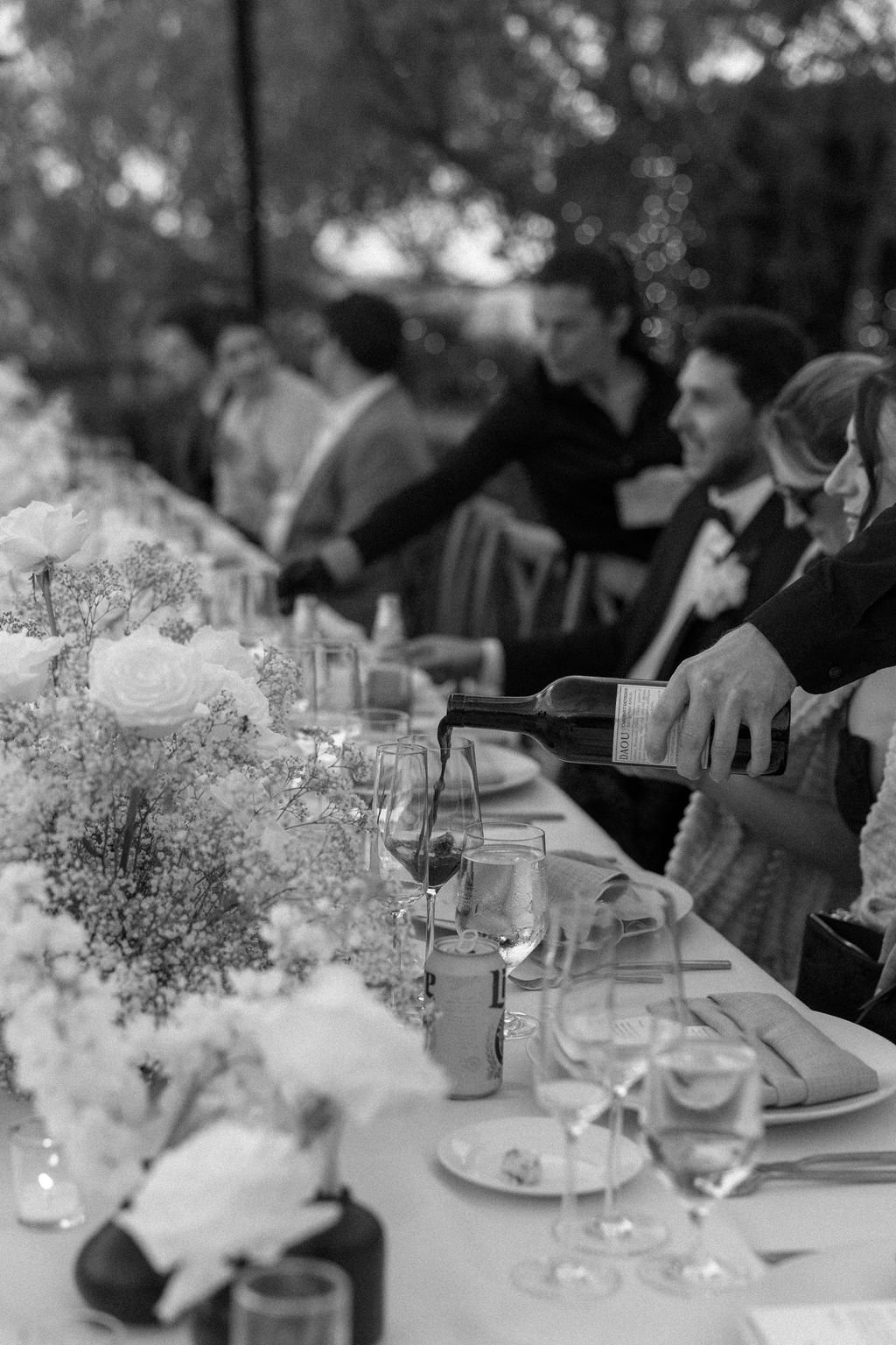 People seated at a long dining table outdoors, with a person pouring wine into glasses, decorated with flowers.