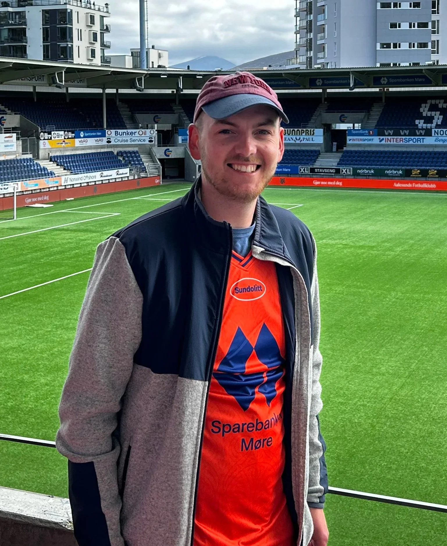 A young man smiling on a football (soccer) field, wearing a baseball cap, a black jacket, and an orange soccer jersey with blue and red accents, with stadium seating and advertisements in the background.