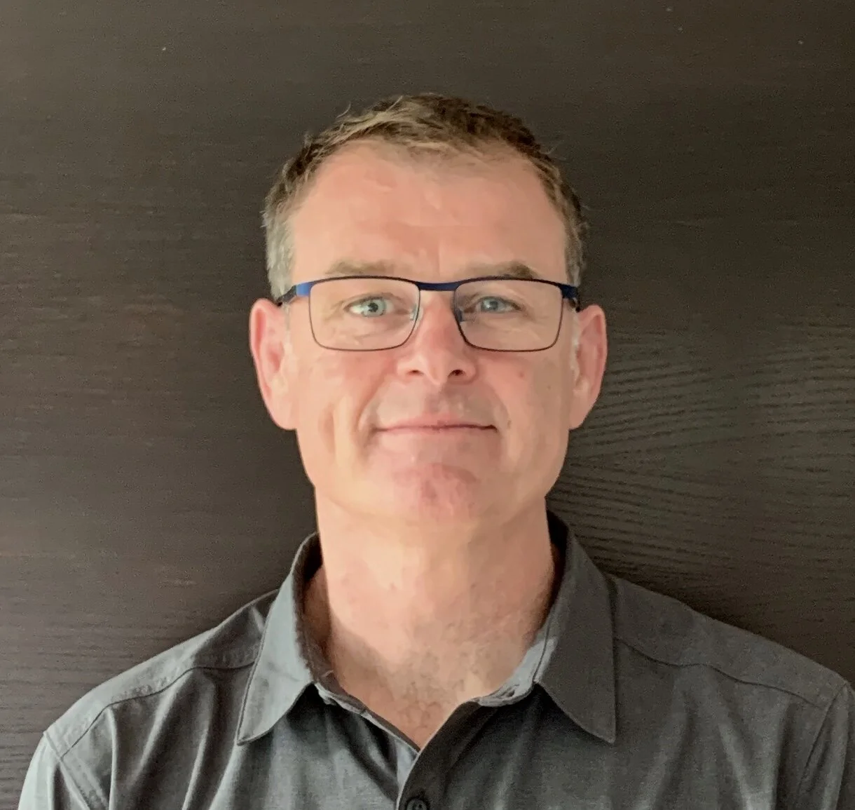 A man wearing glasses and a gray shirt standing against a dark background.